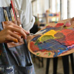 Close-up image of a young creative artist's hands smeared with watercolors holding a spatula mixing color on a palette.